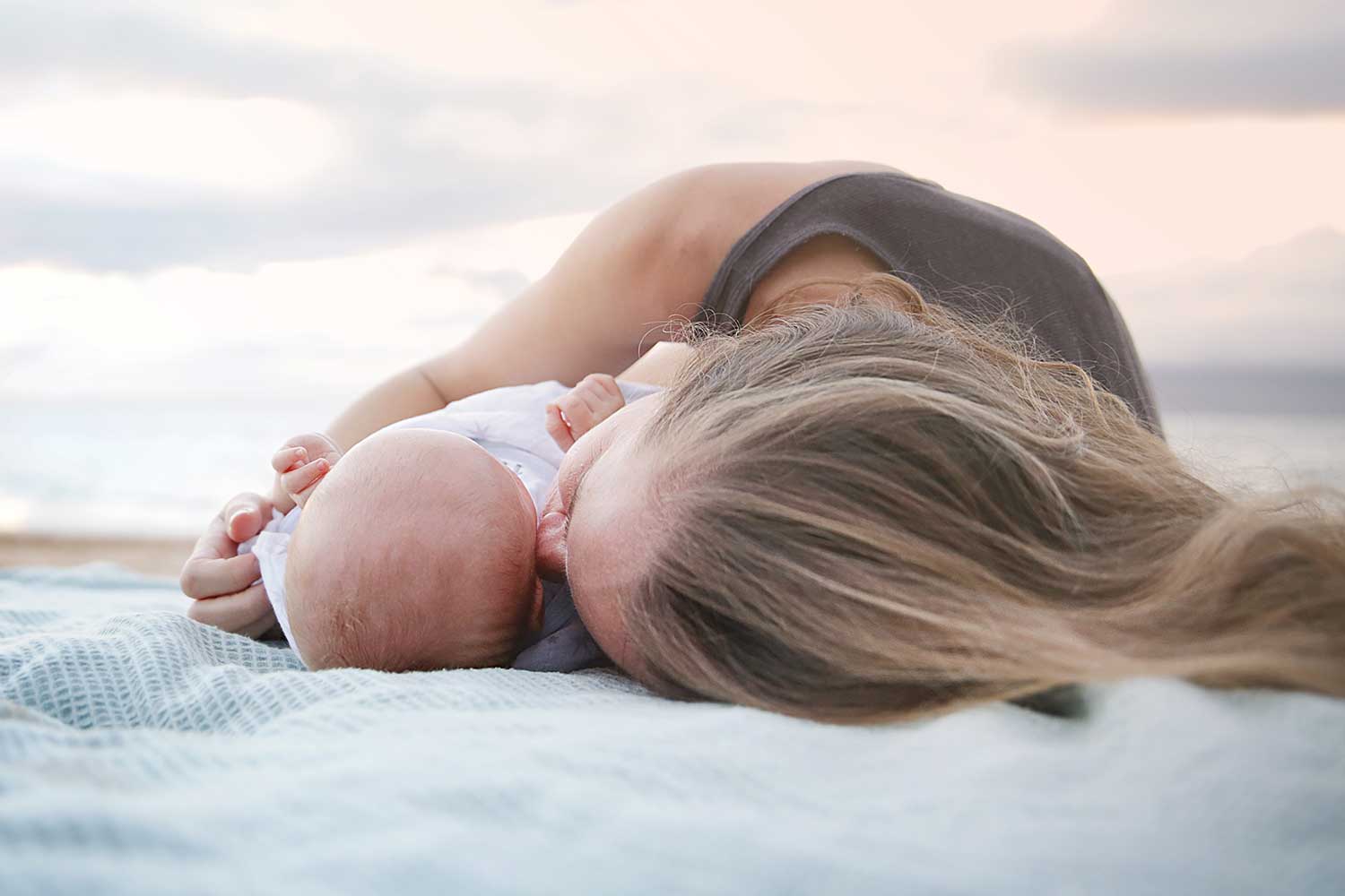 New mom rolls over on beach blanket and snuggles her new baby.