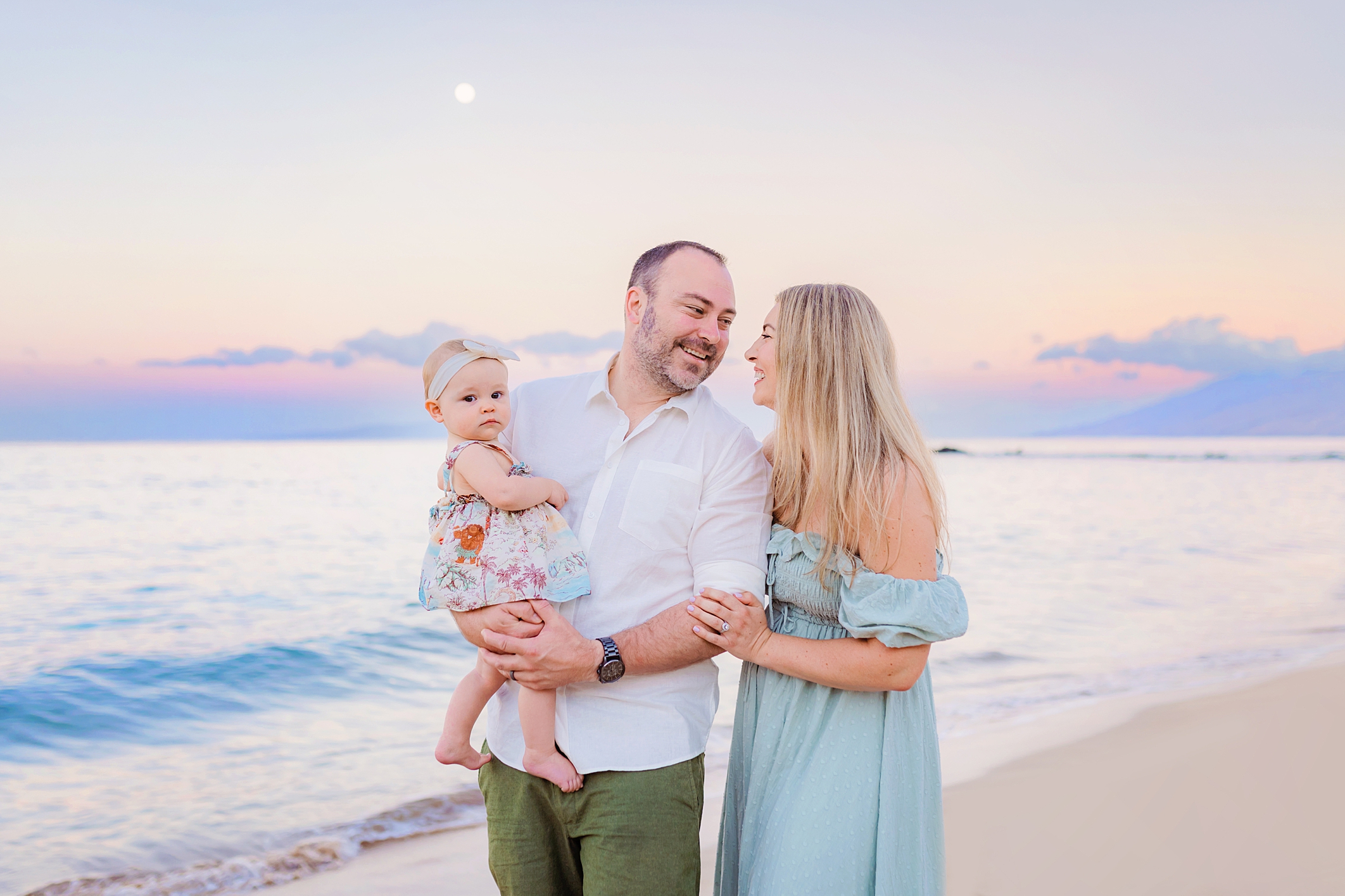 Mom and dad look at one another while holding daughter. toddler stares at the camera while the family is having pictures taken on the beach