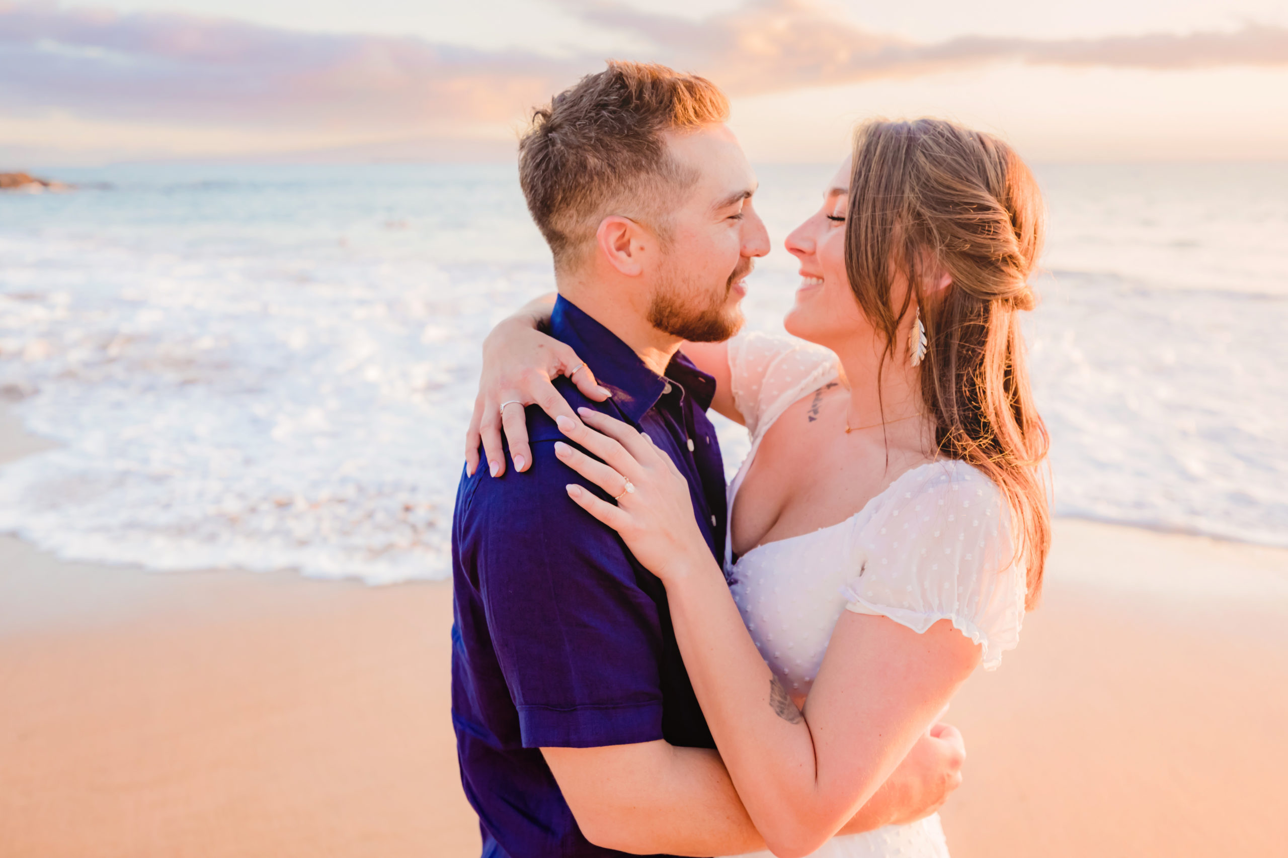 Man in a blue collared shirt holds his new fiance close as they stand on the beach during their proposal photoshoot in Wailea