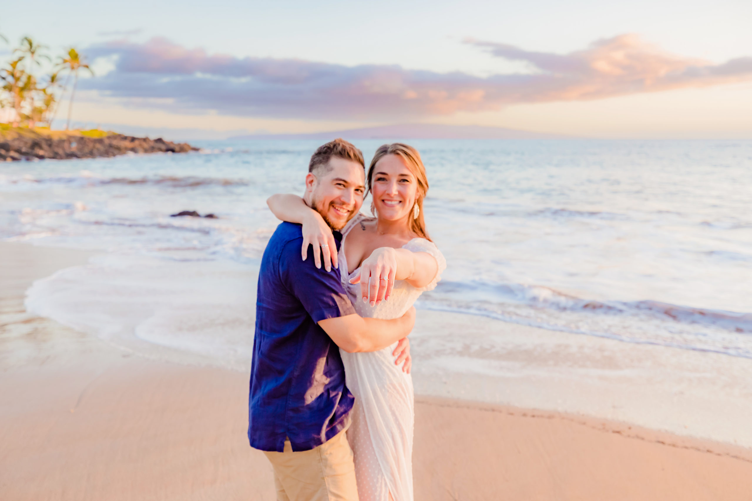 woman in white standing with her new fiance shows off her engagement ring to the photographer at her surprise proposal photoshoot in Wailea
