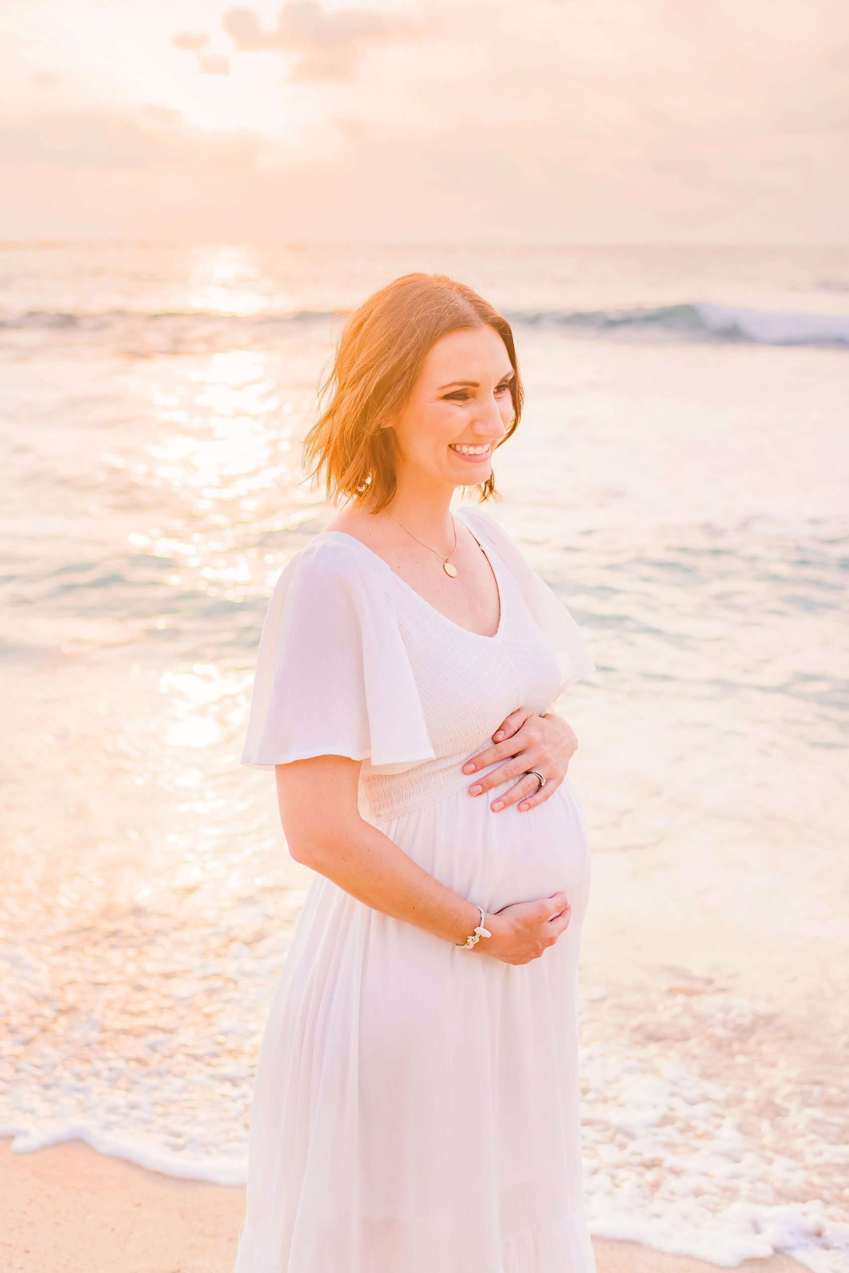 brunette with short hair smiles and looks off camera wearing short sleeved white maternity dress on the beach on the Big Island at sunset