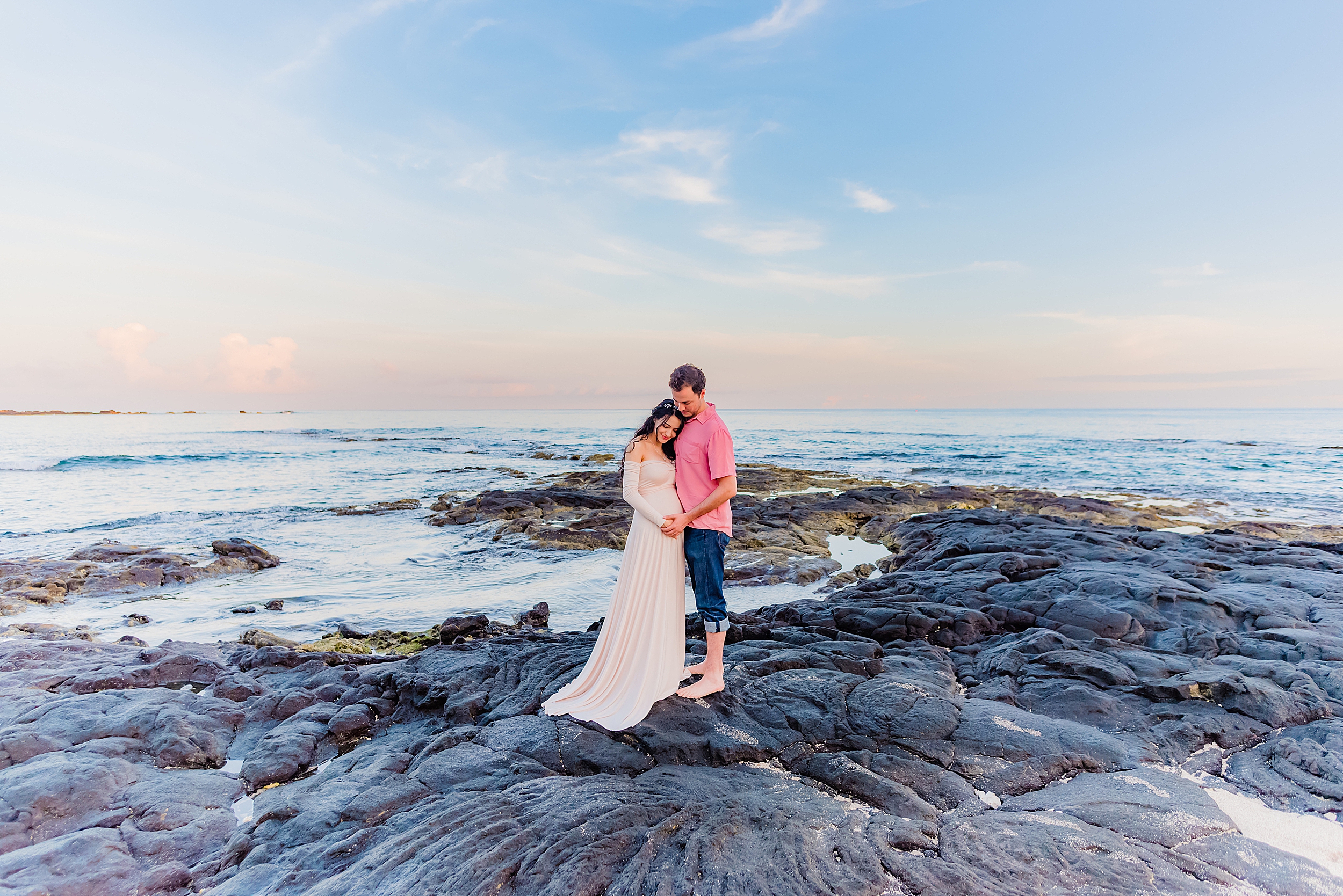 pregnant brunette woman poses in long pink sleeved maternity gown on volcanic rocks during her big island babymoon photography portraits with love + water