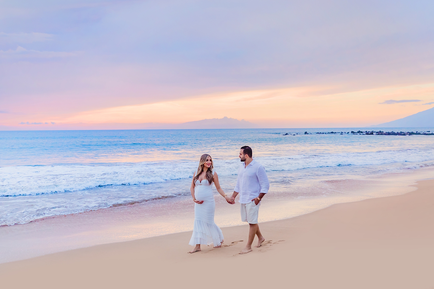 maui babymoon ideas for a pregnant brunette woman with long brown hair and white maternity dress strolls on beach at sunset iwth her husband during a maui photoshoot with love and water photography