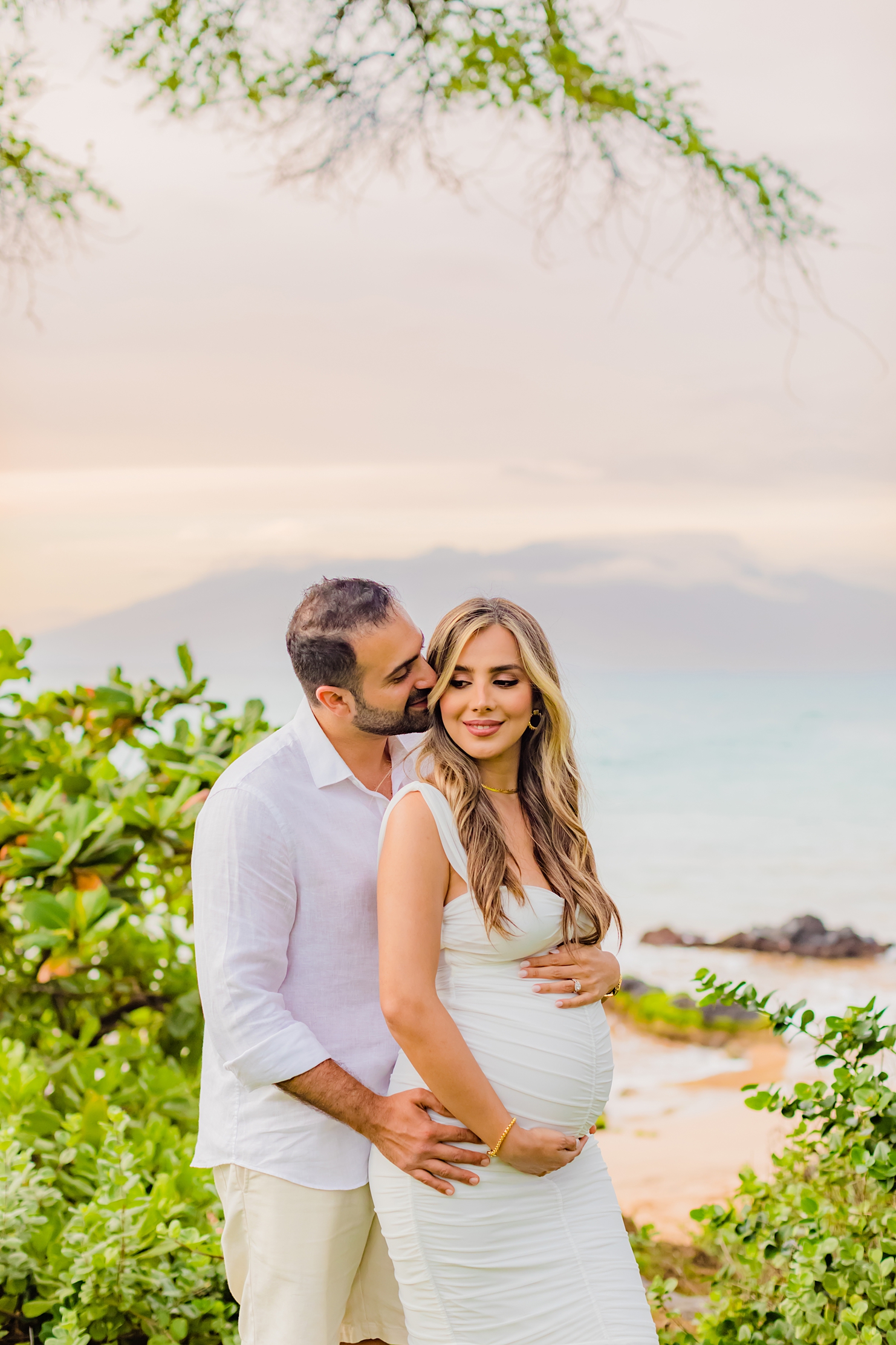 beautiful pregnant woman on maui looks down while husband in white shirt kisses her on the cheek during their babymoon maternity portraits