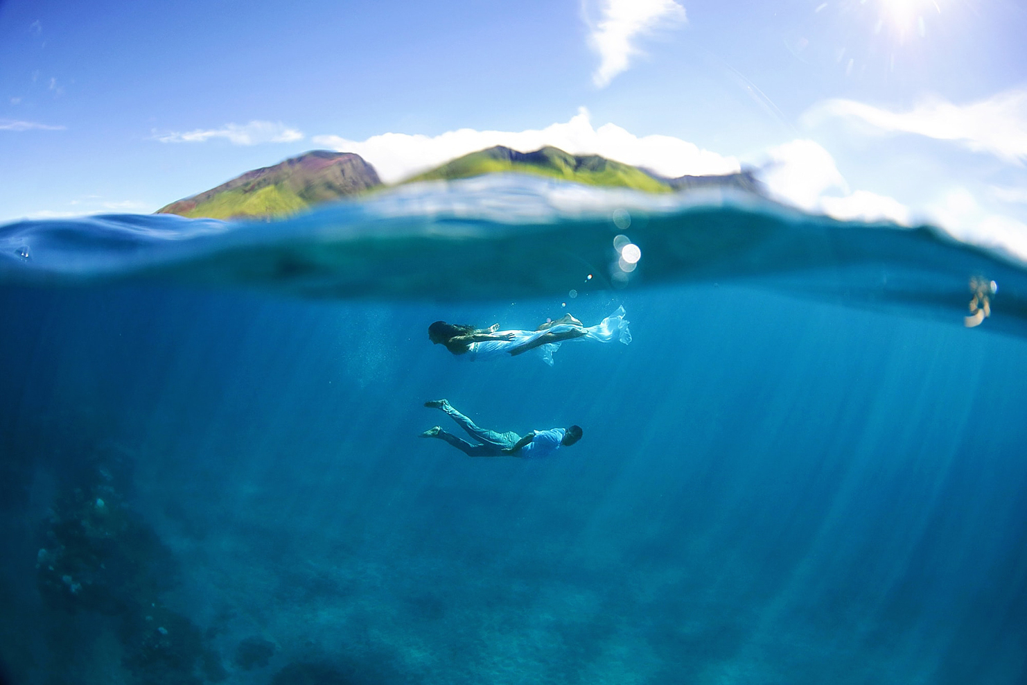 couple underwater and bride with wedding dress swim in the ocean with west maui mountains in the background