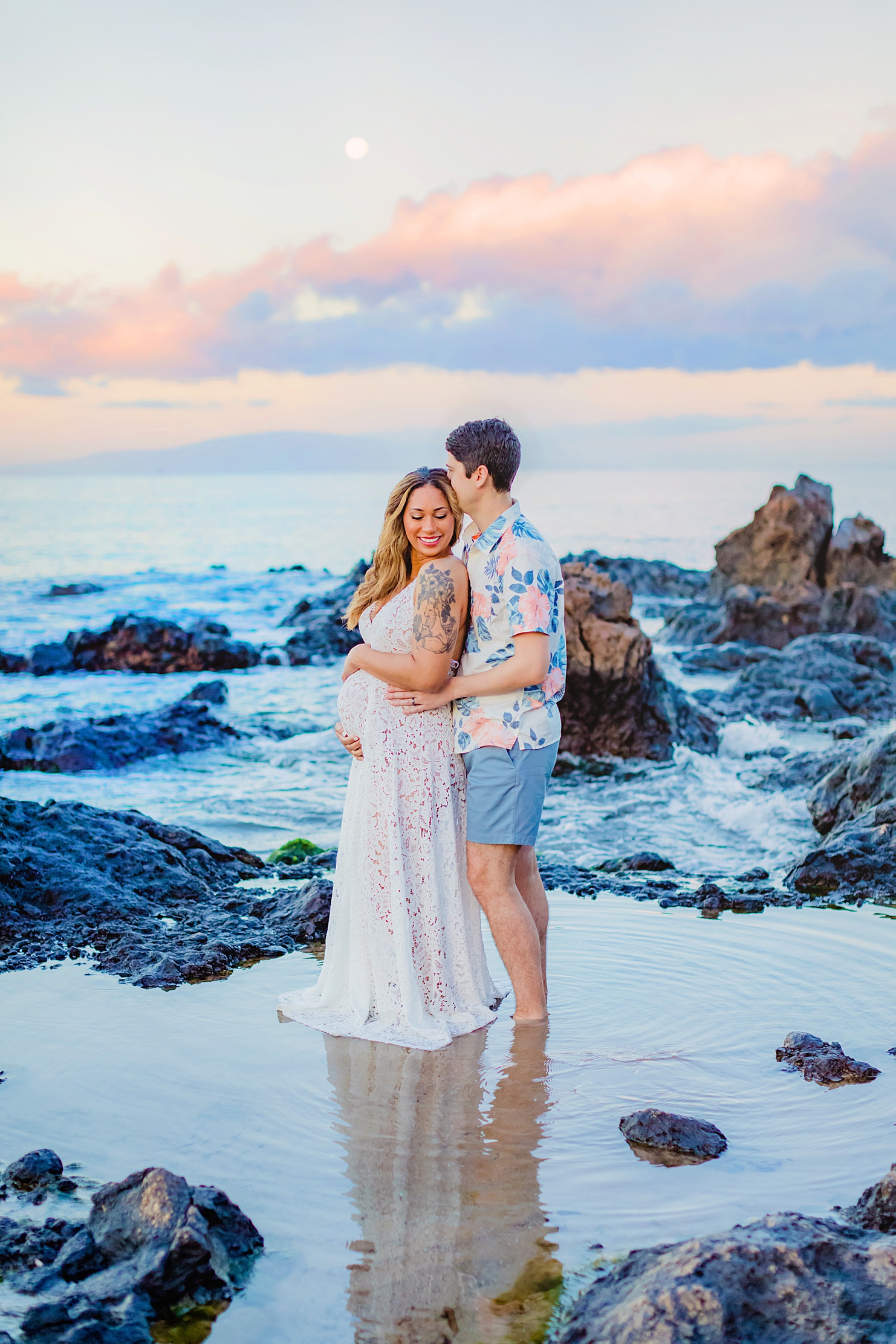 Pregnant woman in white lace gown under the full moon at sunrise in Hawaii