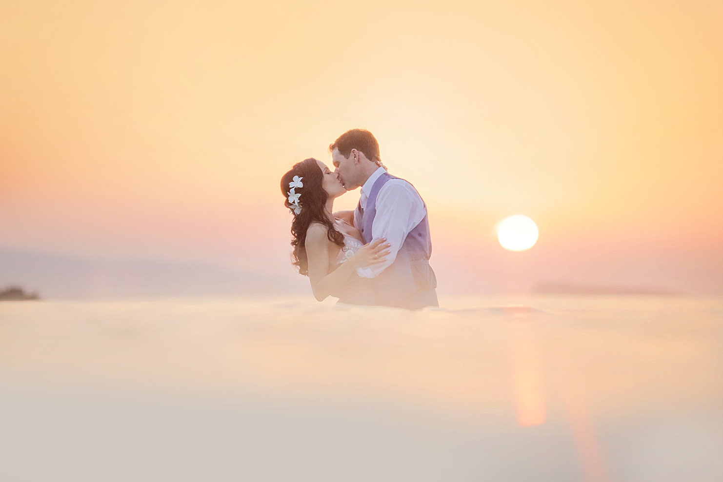woman wearing plumeria flowers in long brown hair kisses her husband in the ocean during sunset in hawaii