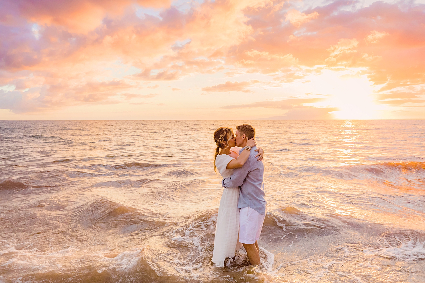 hawaii beach photoshoot ideas couple kisses at sunset on the beach woman with blond hair in a braid wearing a white dress holds her husband around the shoulders