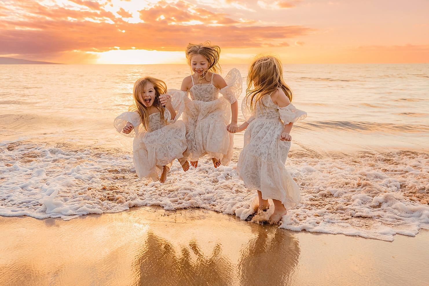 young sisters in white dresses hold hands and splash in the ocean on maui