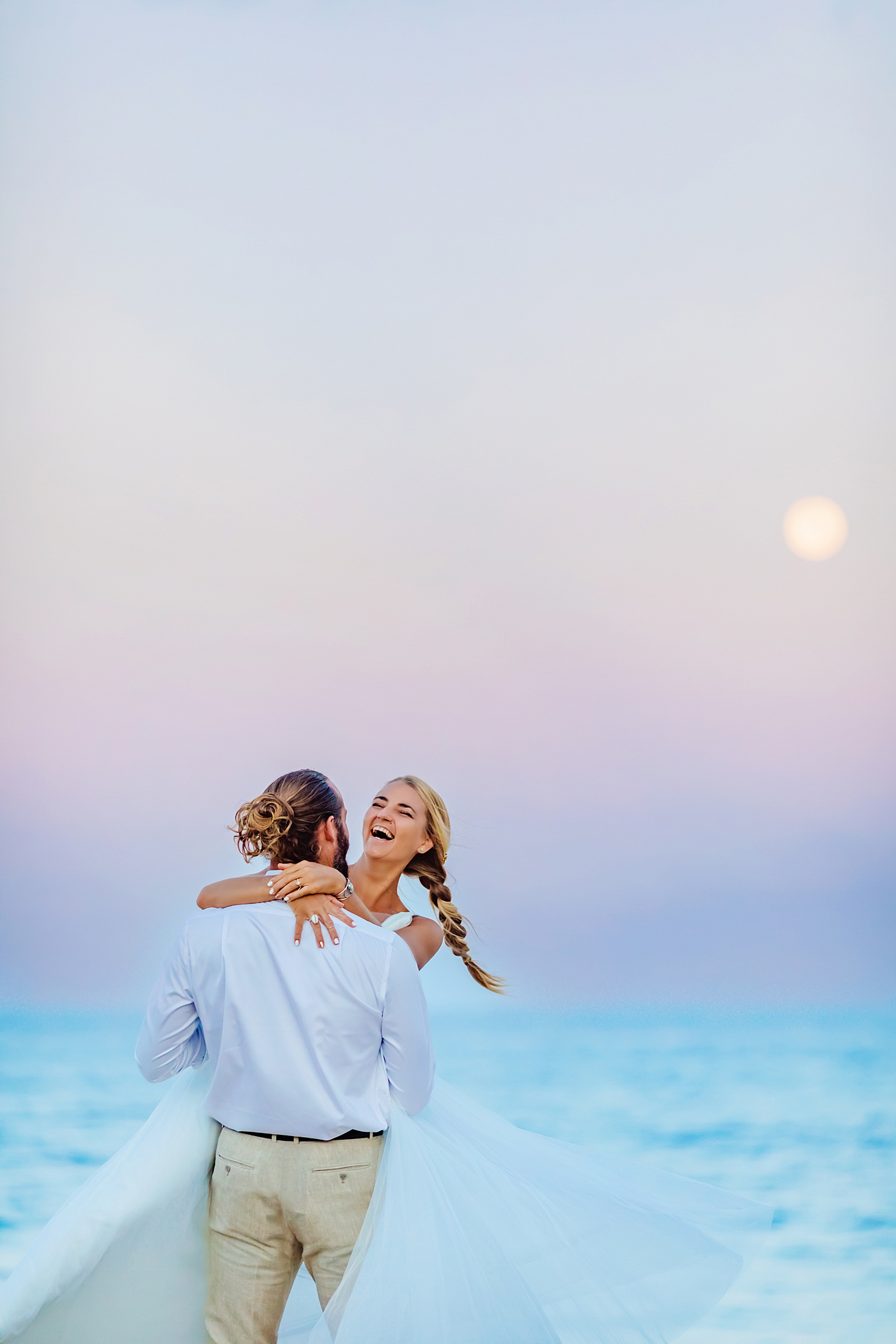 man with white longsleeve shirt twirls wife around under the full moon during a pastel sunrise photoshoot on maui
