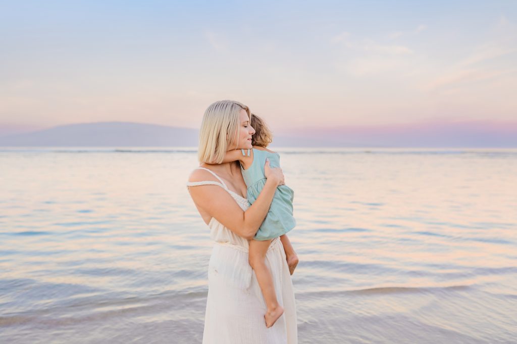 mother with blond hair wearing white cotton gown holds her young daughter and closes her eyes while smiling at the beach during a sunrise photoshoot