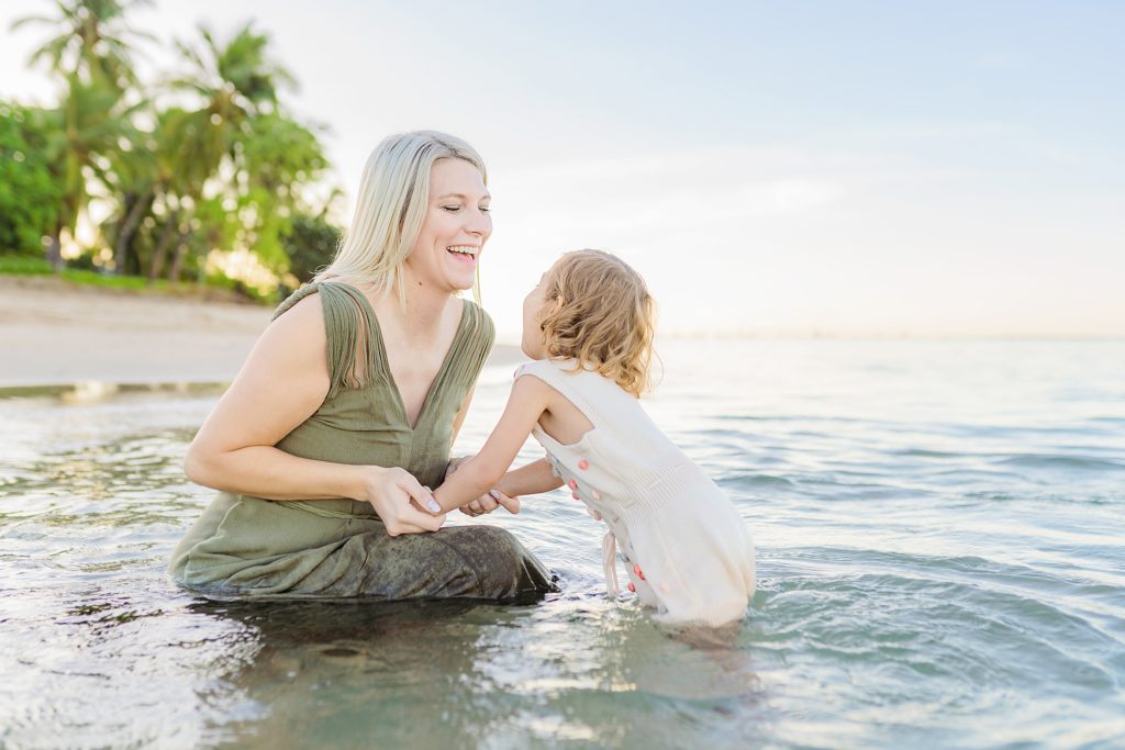 mom with blond hair wearing a hunter green dress kneels in the water with her toddler daughter and splashes around during their lahaina maui family photography session