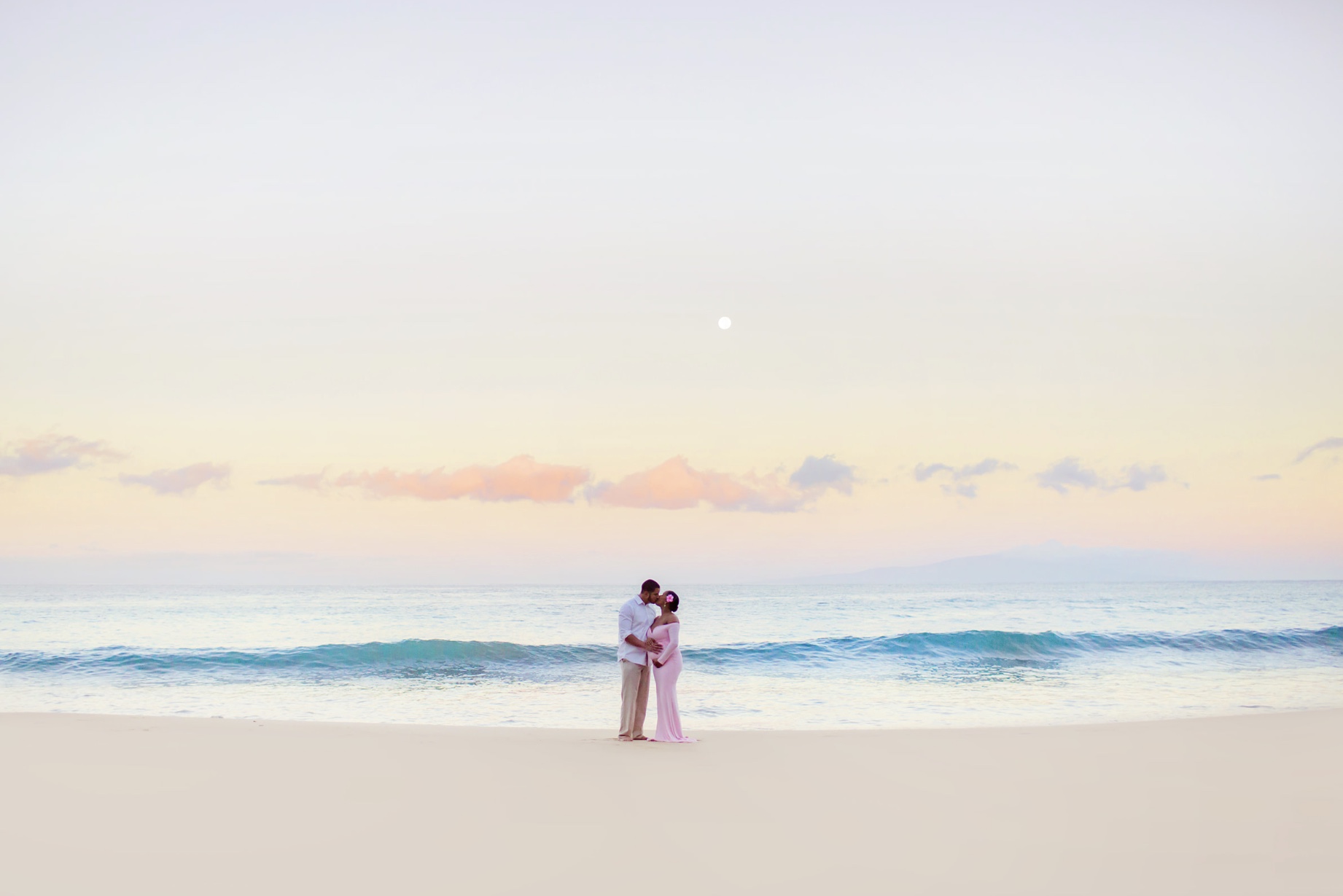 pregnant woman with hibiscus behind her left ear kisses her husband at sunrises during their maui pregnancy announcement