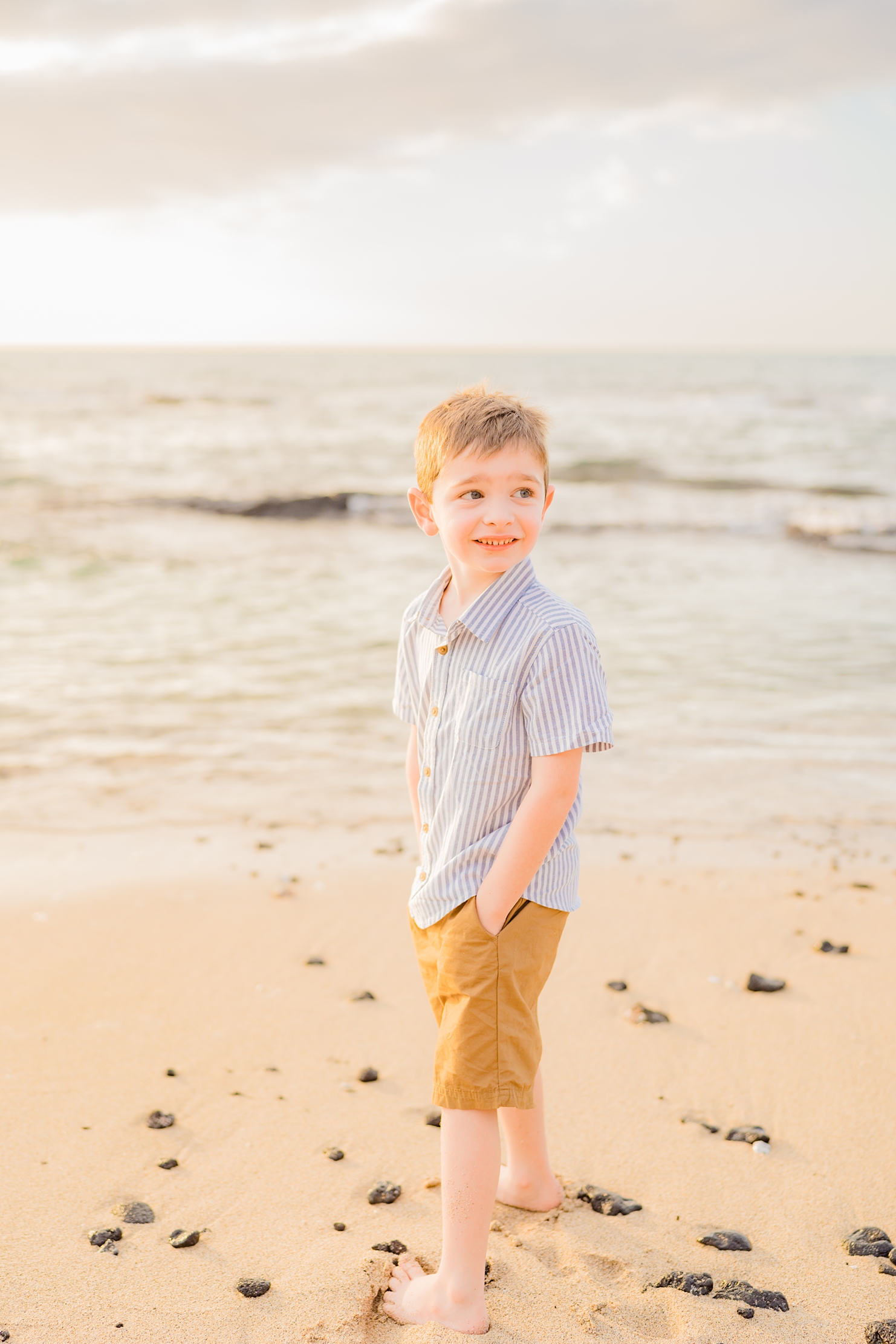 big island family photography session by love and water featuring young boy with hands in his pockets looking off camera at the beach at sunset