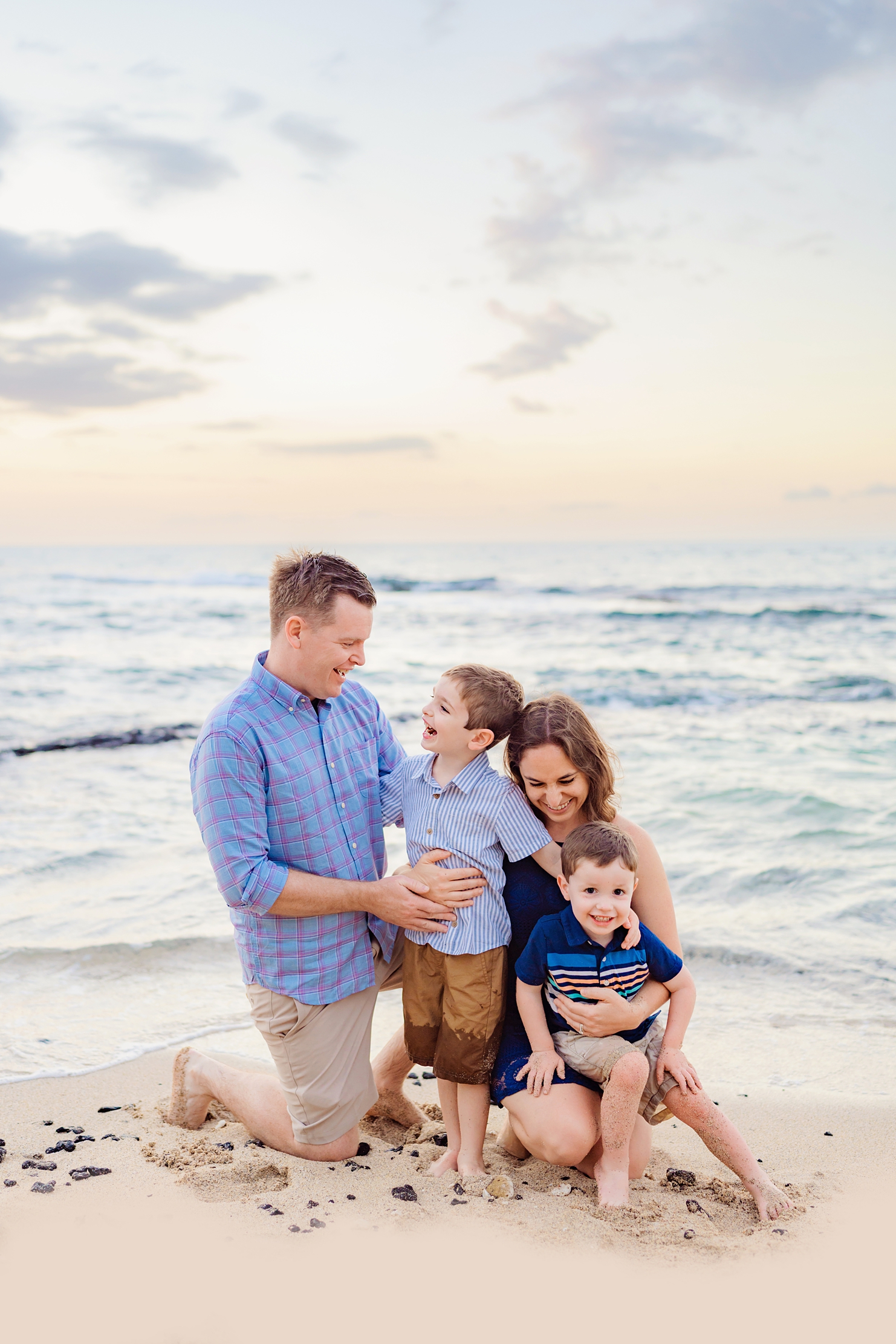 father holds his young son and mother sits in the sand with arms around her youngest boy as the family smiles and laughs on the big island