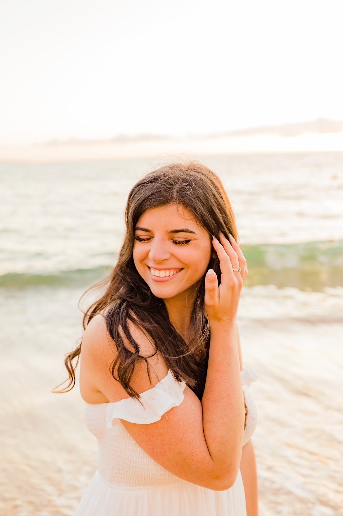 brunette bride smiling and closing eyes while looking off camera at the beach backlit by the setting sun
