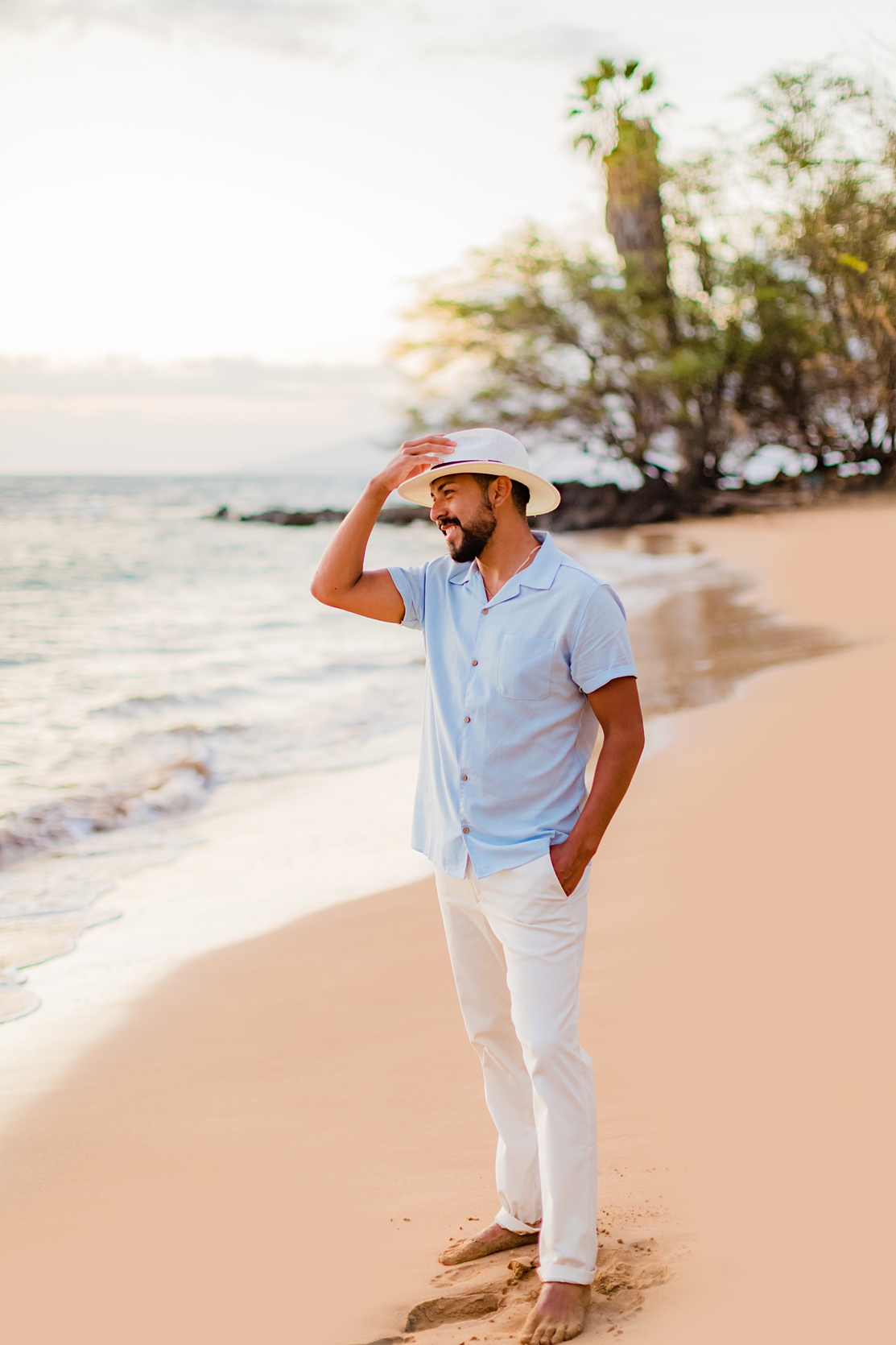 man with white hat and button down short sleeve shirt looks off camera while touching the brim of his hat on the beach