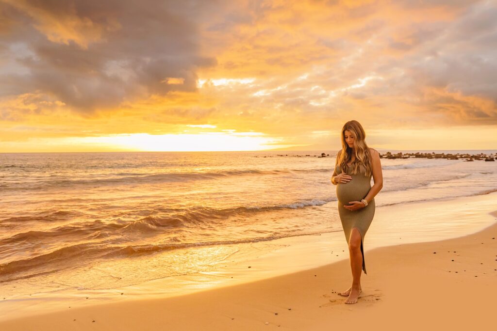 pregnant blond woman wearing green maternity dress looks down at her belly during her maui sunset maternity pictures