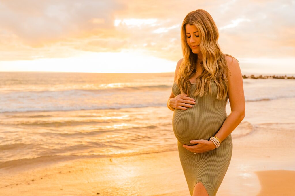blond woman glances down at her pregnant belly and closes her eyes at sunset while posing on the beach in wailea with love and water