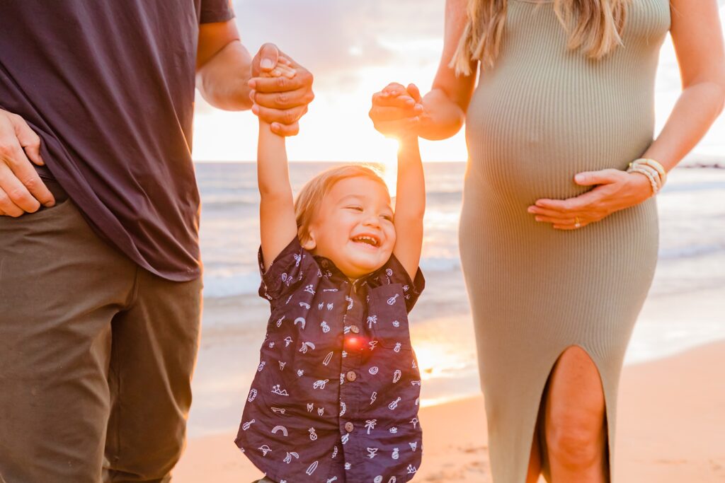toddler smiles and looks off camera as mom and dad lift him up by the arms on the beach during their Maui family photography session