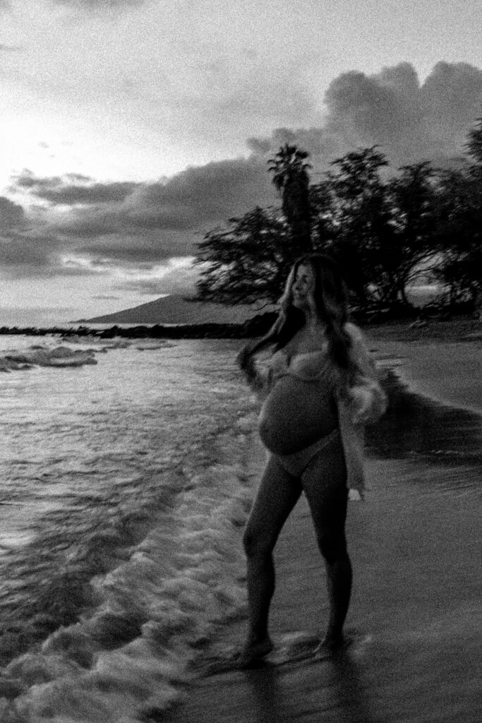 black and white image of pregnant woman holding her long hair walking on a maui beach at sunset