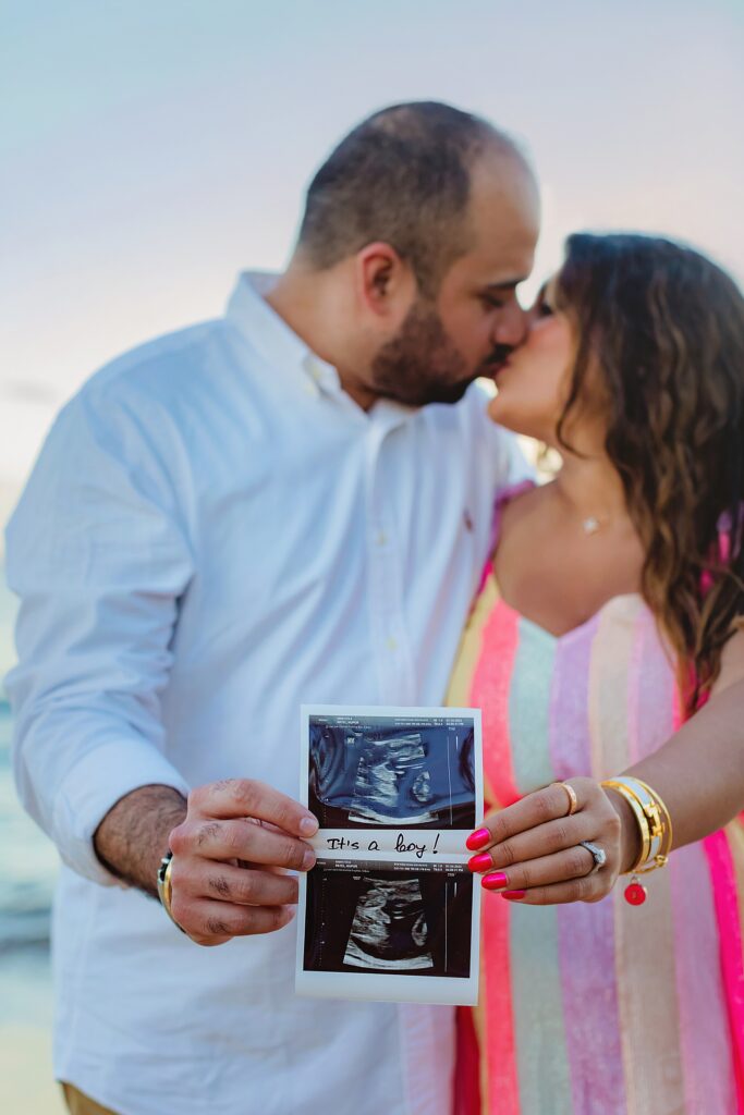 expecting couple holds ultrasound photo to camera while kissing during gender reveal photoshoot