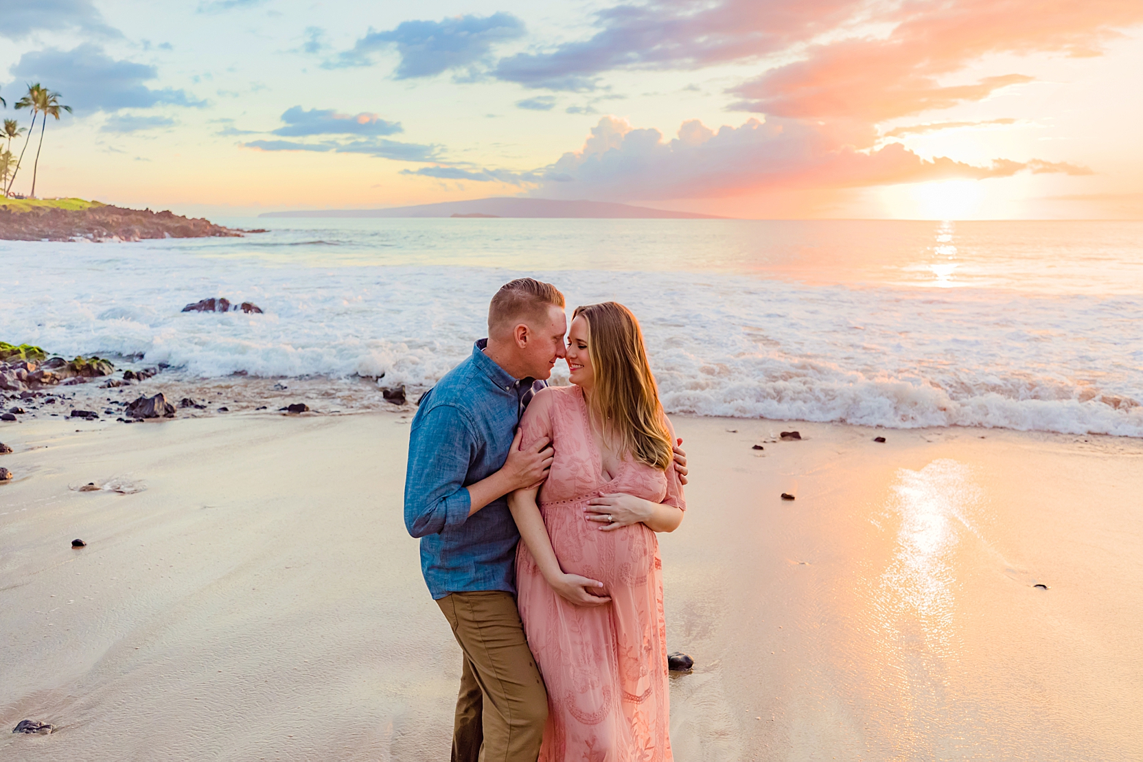woman in pink gown laughs and kisses husband during their maui maternity gender reveal portraits by love and water