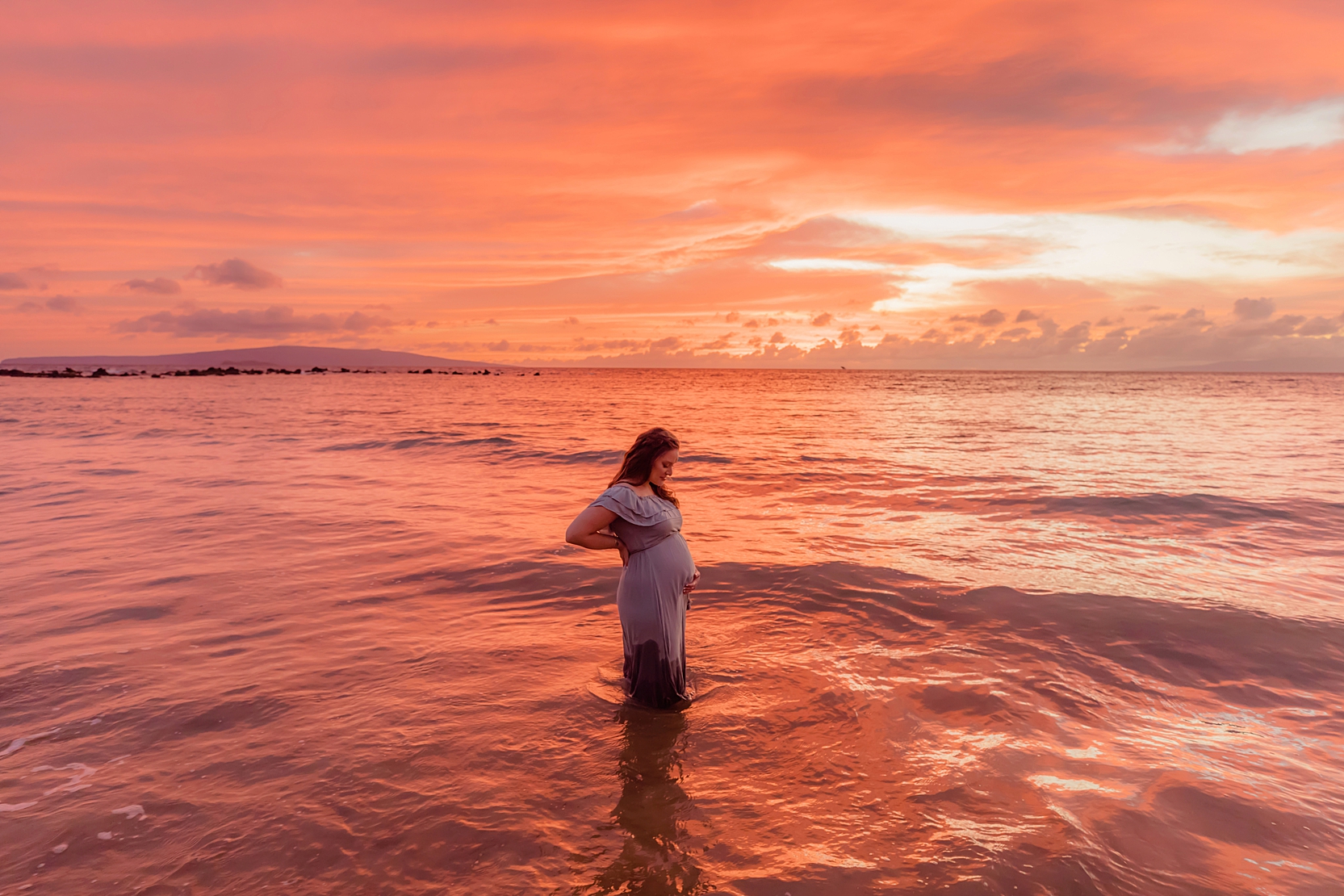 stunning sunset maternity portrait of woman in blue gown knee deep in the ocean looking at pregnant belly