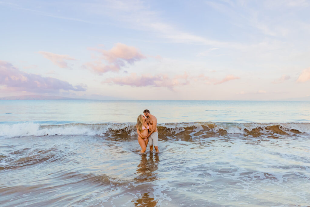 Couple places their hands on woman's growing baby bump while they stand in the water on a beach in Maui