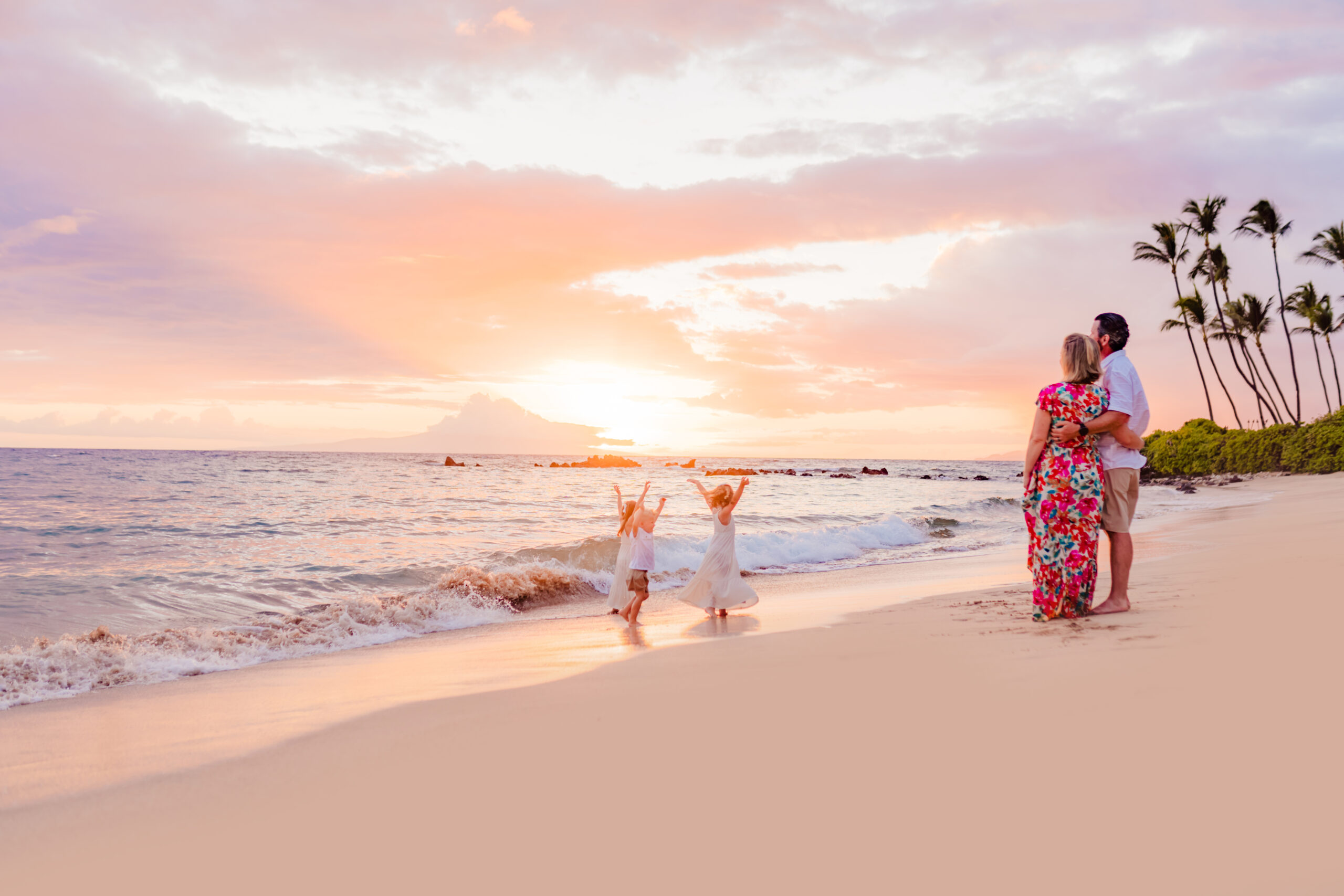 Three kids play on the beach as their parents watch during a picturesque sunset on Maui