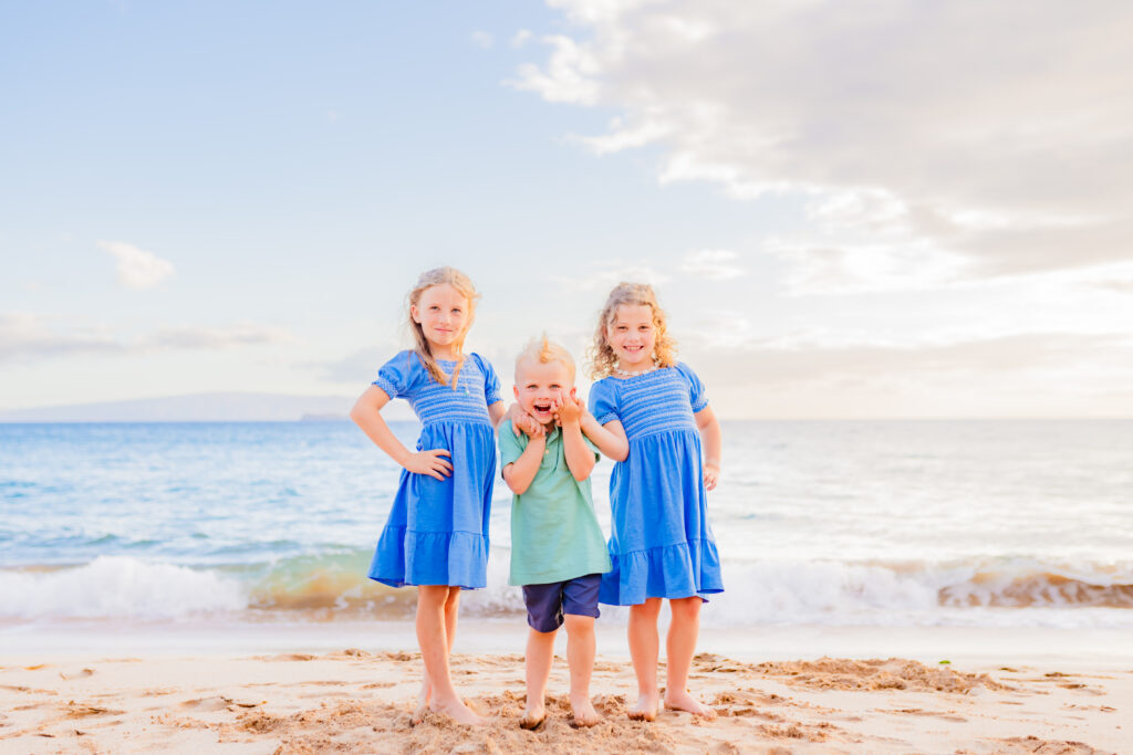 Three kids wearing blue smile for their family photographer on the beach