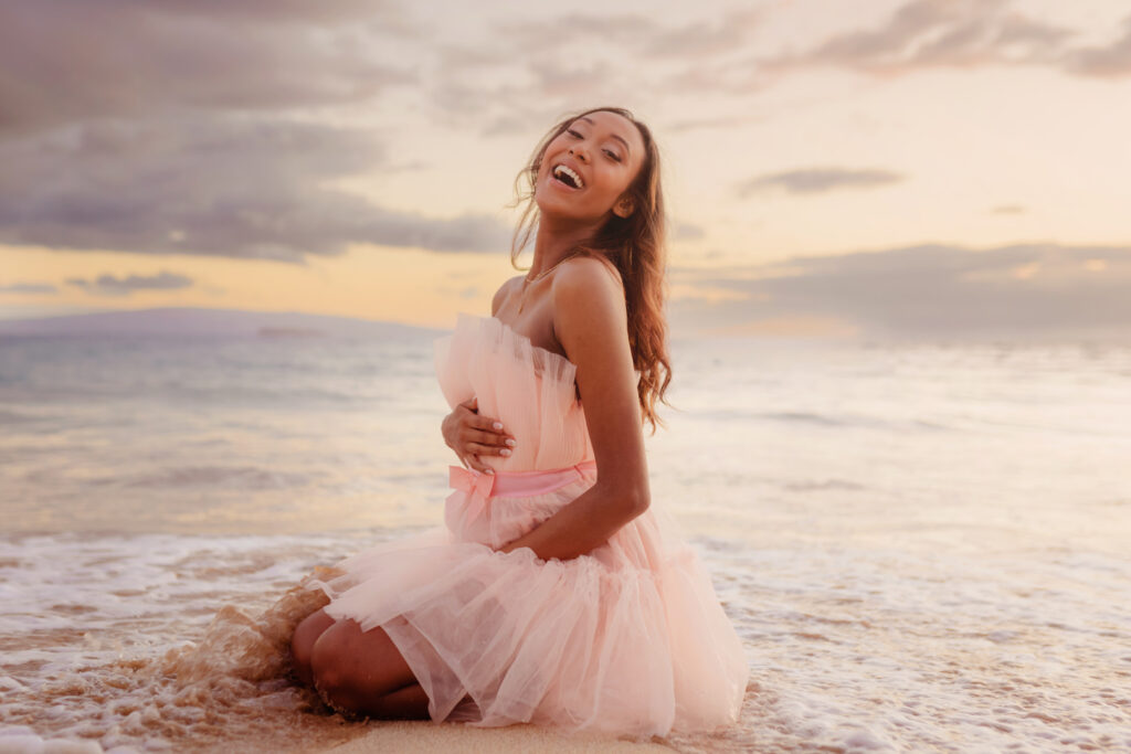 Woman sits on the beach and poses in a pink tulle dress for maternity photos on Maui