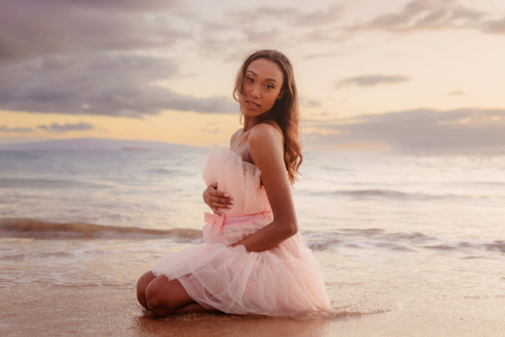 Woman poses on the beach in a pink tulle dress for maternity photos