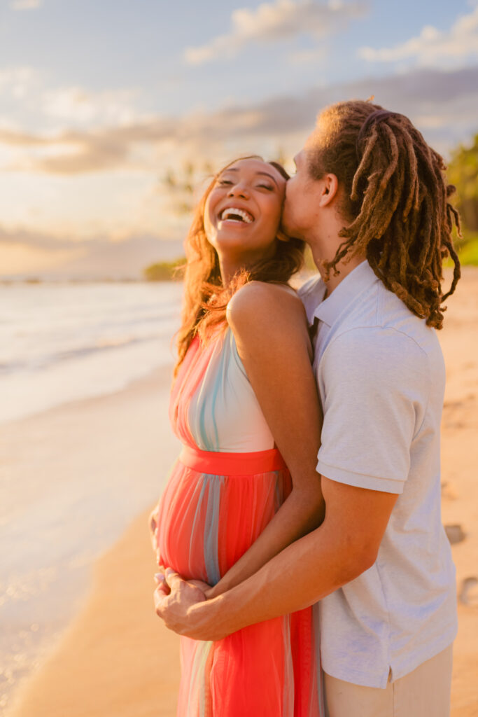Man plants a kiss on his pregnant partner as she smiles enjoying the sunset at the beach