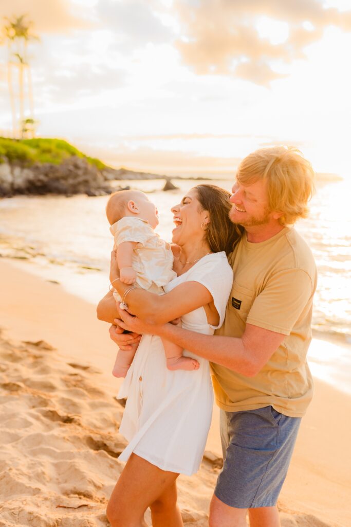 Brunette mom wearing white dress smiles and laughs while husband holds her and they stare at infant on the beach