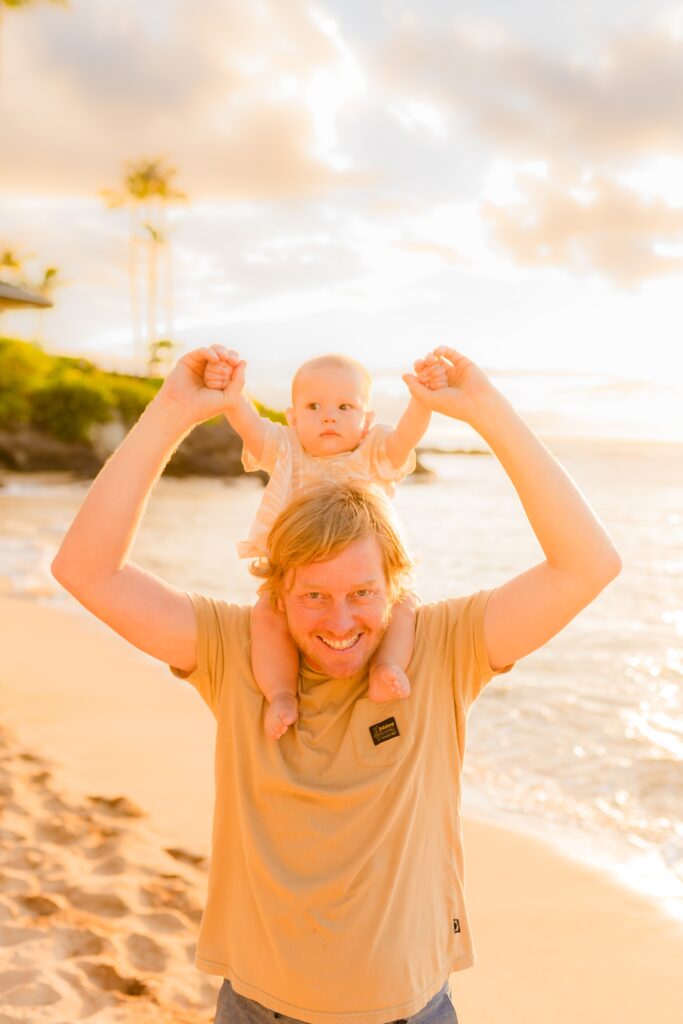 Infant looks unsure on dad's shoulders holding up and smiling at camera on Maui during Love and Water photoshoot in Kapalua Bay