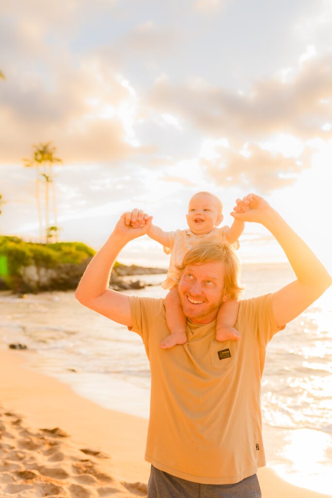 Dad looks up at young son on his shoulders during Maui family photography session in Kapalua Bay