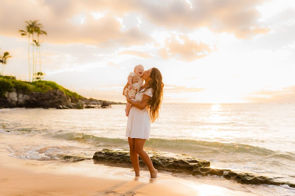 Mother in white dress holds her infant son on the shoreline at Kapalua Bay, Maui and kisses him on the cheek at sunset