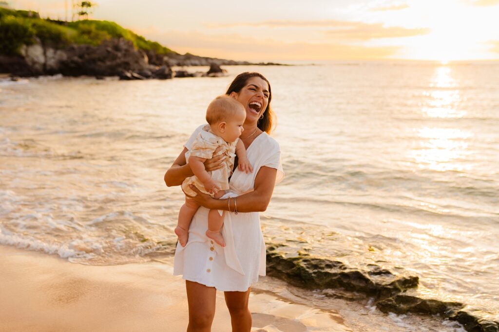Mom laughs off camera wearing white dress holding infant son during photoshoot on Maui