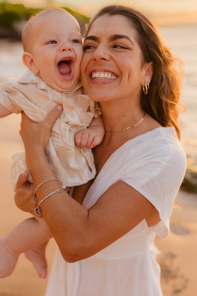 Young brunette mother holds her young son and smiles up at him on Maui as he laughs off camera at dad
