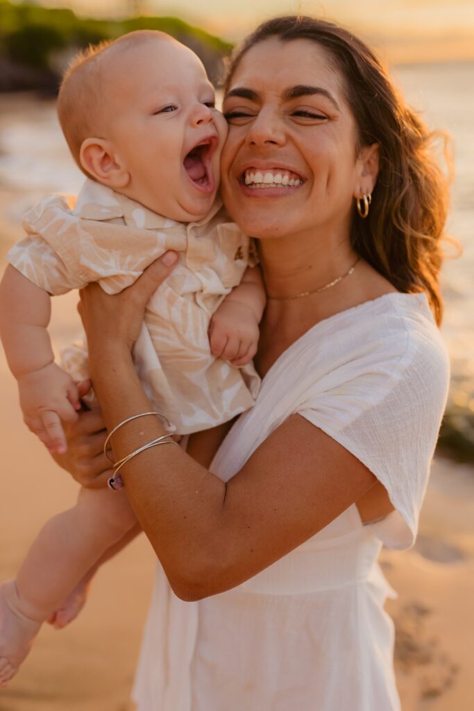 Infant boy smiles and laughs as he reaches for his mother's cheek during sunset family photoshoot on Maui