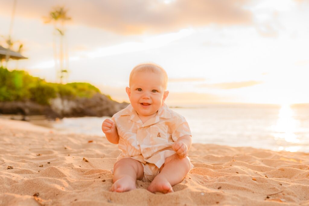 Infant wearing beige beach outfit smiles and sits on the sand in Kapalua Bay