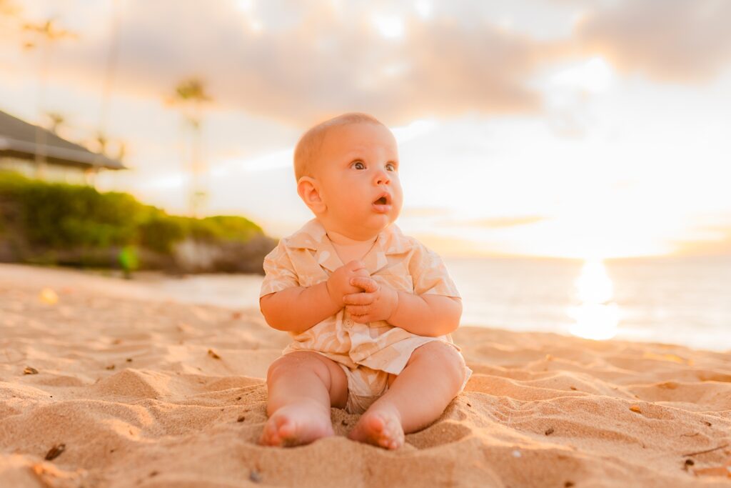 Infant looks off camera while sitting wearing a cute beach outfit and playing with sand