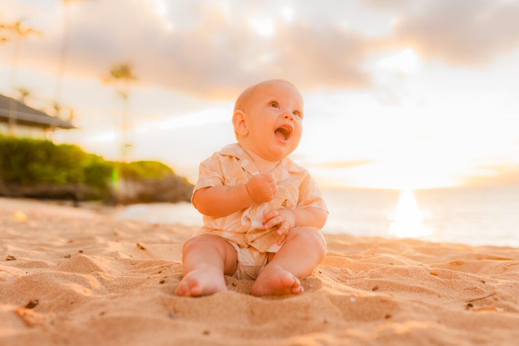 Young boy sits on the sand and looks up at parents during sunset photoshoot in Kapalua Bay