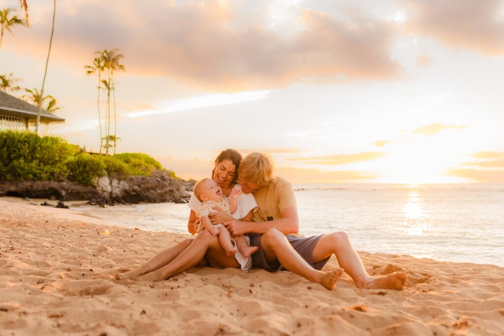 Young boy sits in mother's lap in the sand at sunset and laughs at parents gleefully