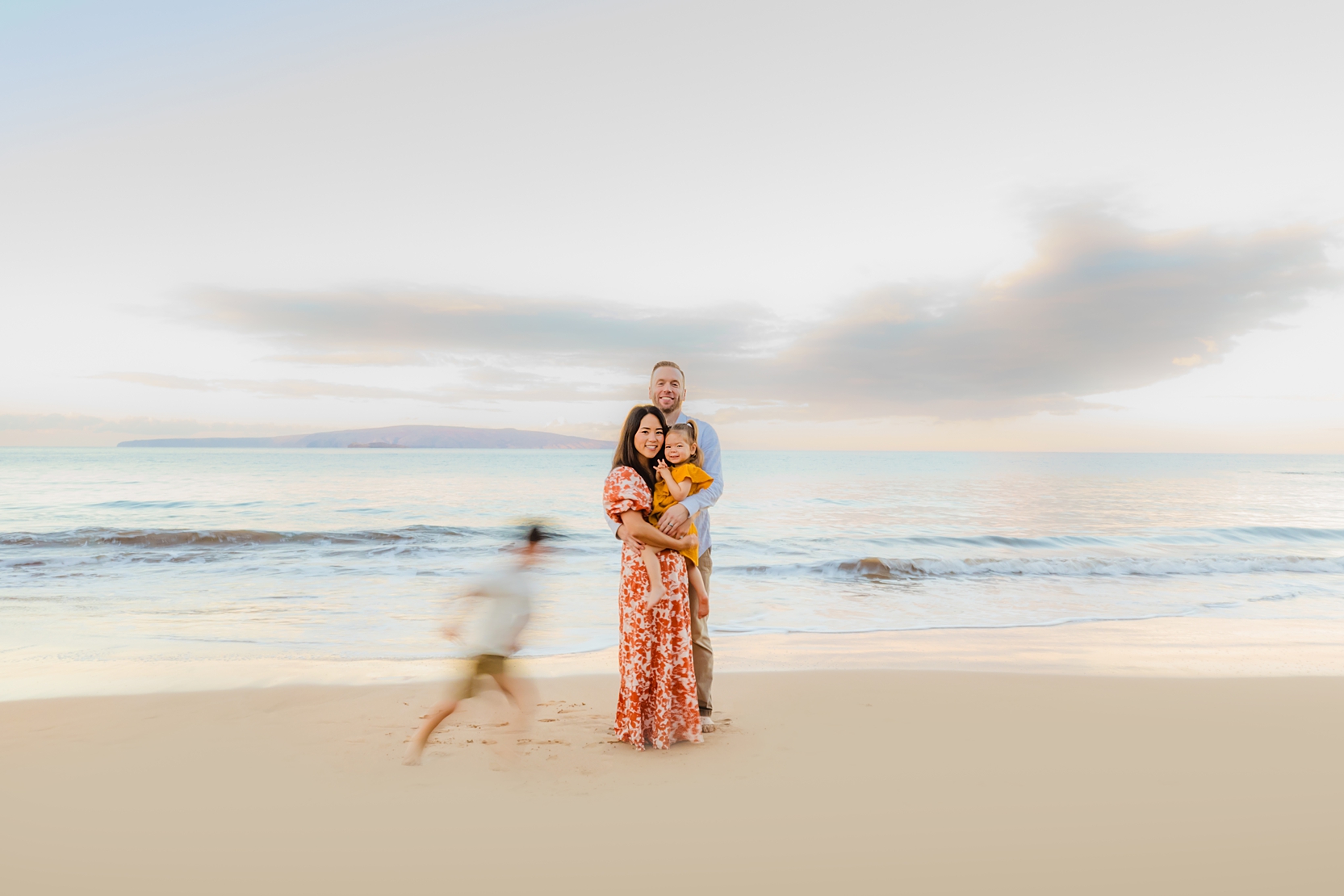 artistic maui family pictures featuring a young family wearing organce and yellow staring at the camera while young son circles them running