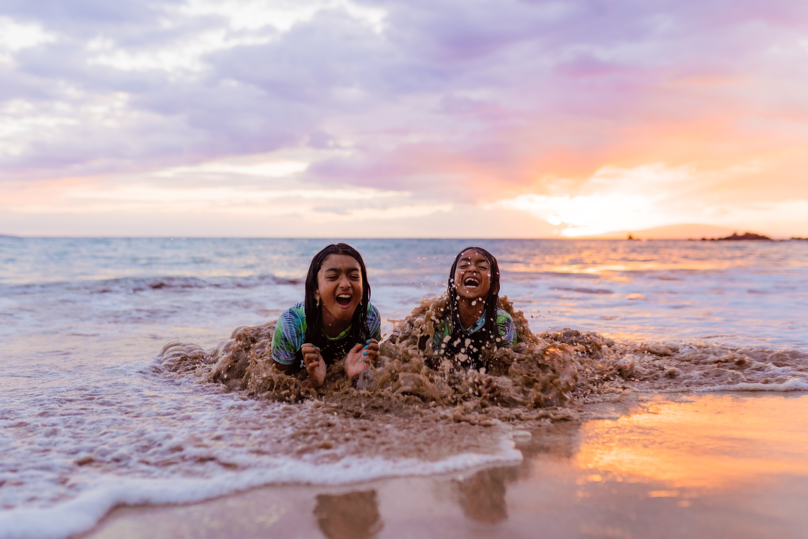 fun maui family photoshoot in wailea with two young girls laying in the sand smiling and laughing as a wave splashes them unexpectedly