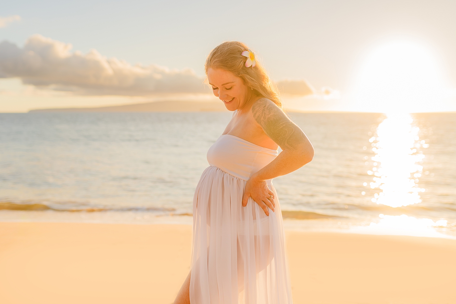 Woman in sheer white maternity gown places hands on hips and smiles down at her pregnant belly on the beach in hawaii