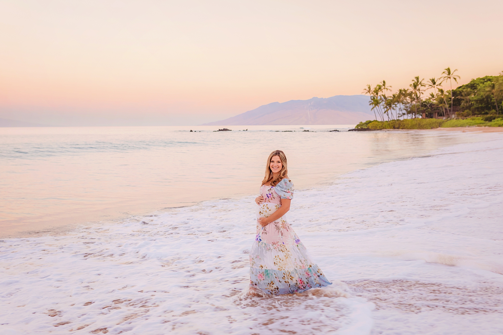 Pregnant woman holds her belly and stands in the water while waves wash ashore at sunrise in Wailea