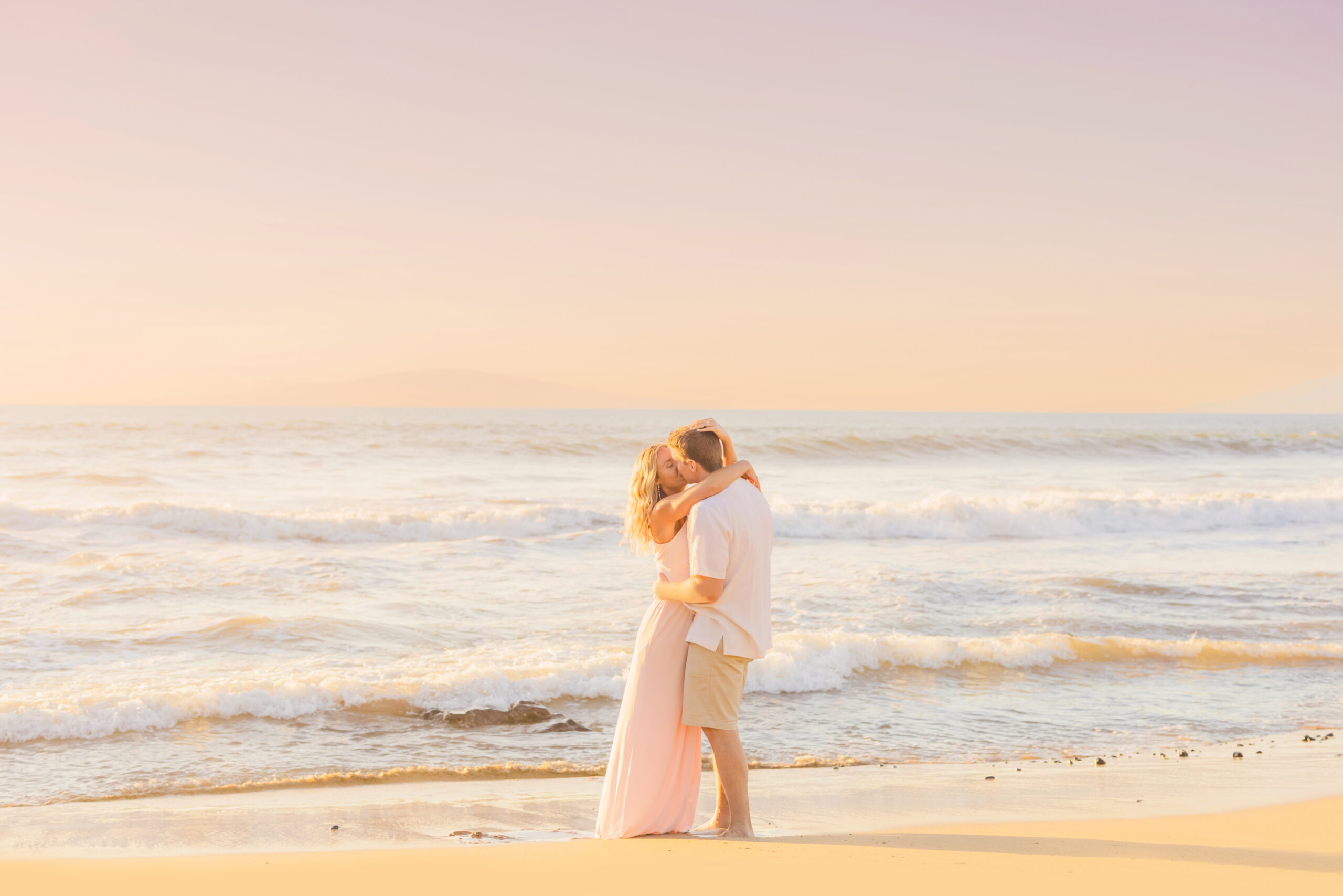 male and female couple kisses on a beach in Hawaii as the waves crash on the shore during an engagement photoshoot